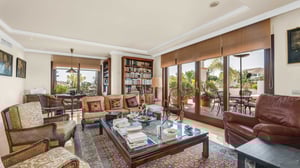 Elegant living room with a cozy atmosphere, featuring classic armchairs, a large bookshelf, glass coffee table filled with books, and large sliding doors opening onto a lush terrace.