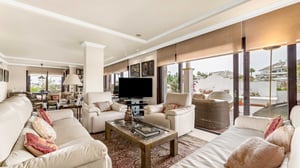 Spacious living area with cream leather sofas, a large TV, decorative pillows, and floor-to-ceiling windows that flood the space with natural light and offer access to a sunny terrace.