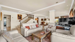 Wide-angle view of the living room showcasing multiple sitting areas, traditional furnishings, artwork on the walls, and a staircase leading to the upper floor.