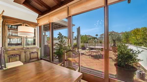 Dining room with rustic wooden furniture, a vintage hutch, and large glass doors opening onto a sun-drenched terrace filled with greenery.