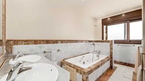 Elegant bathroom with double sinks, a built-in whirlpool bathtub, and marble finishes in warm tones, complemented by a large frosted window.