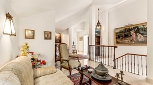 Elegant upper-level sitting area with vintage armchairs, a floral-patterned sofa, and antique decor. Features classic artwork, a carved handrail overlooking a staircase, and a decorative hanging lantern.