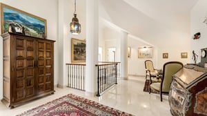 Refined landing area with white pillars, oriental rugs, a large wooden wardrobe, and a variety of paintings. The space is well-lit and blends traditional furnishings with a clean, airy layout.