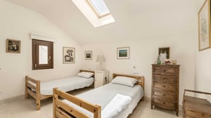 Bright bedroom with two single wooden beds, simple white bedding, a skylight, and minimalist artwork. Includes a wooden chest of drawers and small window for additional natural light.