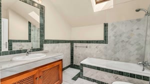 Bathroom with elegant green and white marble tiling, bathtub with overhead shower, large vanity mirror, and a skylight offering natural brightness.
