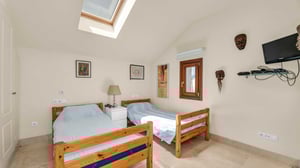 Cozy twin bedroom with two wooden beds, skylight, and eclectic wall art including tribal masks. Includes a small TV mounted on the wall and a simple white nightstand.