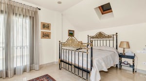 Traditional-style bedroom with a pair of ornate wrought-iron beds, sheer floor-length curtains, and antique decor. Natural light filters through a skylight and balcony door.