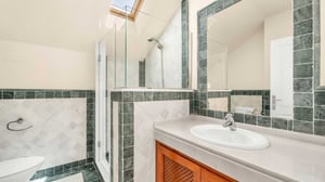 Modern bathroom with green and white tiled walls, glass shower enclosure, and large mirror. The room is well-lit by a skylight above the shower area.