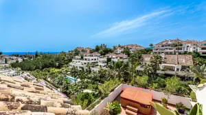 Panoramic view of a lush Mediterranean residential complex with terracotta rooftops, palm trees, and distant sea views under a bright blue sky.
