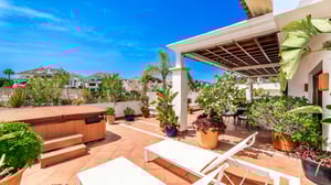 Outdoor relaxation area with a covered terrace, hot tub, sunbeds, and potted plants, surrounded by views of nearby white buildings and blue sky.