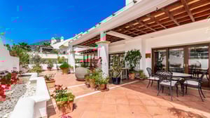 Covered terrace with wrought iron outdoor dining set, surrounded by Mediterranean flora and flowers, with mountain views in the background.