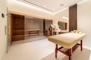 Luxury spa room with a massage table, large round mirror, wooden vanity, and a glass-front sauna with warm wooden interiors.