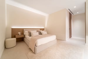 Minimalist bedroom in neutral tones with a padded headboard, floating bedside table, soft lighting, and a cozy carpet.