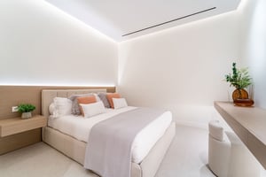 Bright bedroom with soft white and beige decor, accented with orange pillows, and a sleek floating desk with a potted plant.