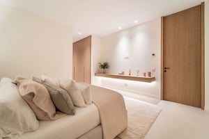 Serene bedroom with layered neutral-toned pillows, soft lighting, a floating shelf, and built-in closets for a clean look.