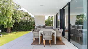 Covered outdoor dining area with a woven chair set and rectangular table, surrounded by a lush garden and outdoor kitchen.
