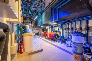 Utility and maintenance room with exposed blue piping, water tanks, and technical equipment, brightly lit and organized.
