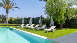 Tranquil garden with sun loungers and umbrellas alongside a modern swimming pool, surrounded by tall greenery and a palm tree.