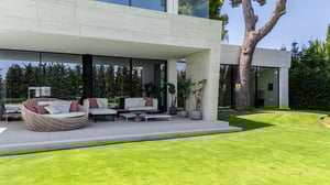 Elegant outdoor lounge with contemporary sofas and decorative plants, under a covered terrace connected to a modern villa.