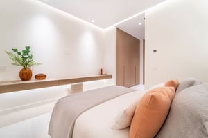 Minimalist bedroom with soft lighting, floating shelf with decorative items, neutral tones, and accent pillows in soft hues.