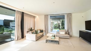 Spacious modern living room with large floor-to-ceiling windows, two beige sofas, a low coffee table, and garden views with a pool.