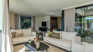 Living area with two beige sofas, black matte coffee tables, full-height curtains, and views of a private garden through glass walls.