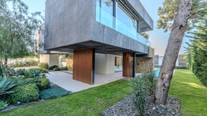 Modern villa exterior with grey stone cladding, vertical wood paneling, and a landscaped garden with lush tropical plants.