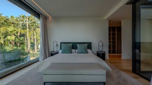Master bedroom with green headboard, decorative pillows, light bedspread, wide windows overlooking palm trees, and wooden flooring.