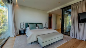 Side view of the same bedroom featuring a bench at the foot of the bed, beige curtains, and access to a glass-walled terrace.