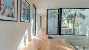 Hallway with light wooden floors, white walls decorated with colorful framed art, and large windows overlooking an internal garden with trees.