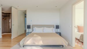 Minimalist bedroom with a beige upholstered bed, walk-in closet, open door to adjacent bedroom, and wood flooring.
