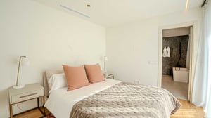 Bright bedroom with soft neutral tones, pink accent pillows, and direct access to an ensuite bathroom with a freestanding tub.