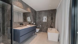 Elegant bathroom with double vanity, freestanding bathtub, black fixtures, grey marble-effect walls, and natural light from tall windows.