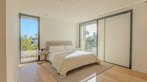 Sunlit bedroom with light bedding, large windows on two sides, and direct access to a balcony with garden views.