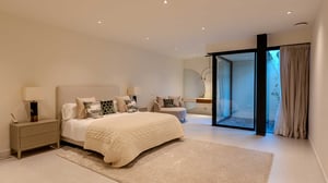 Spacious bedroom with neutral decor, designer pillows, lounge chair, vanity area, and a glass wall looking onto a small patio.