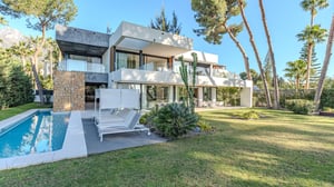 Rear exterior view of a luxury villa with geometric architecture, stone and concrete finishes, floor-to-ceiling windows, and lush garden with pool.