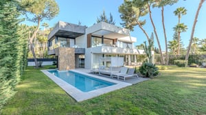 Angled view of a contemporary villa with lap pool, private garden, and shaded lounge area set amid tall pine and palm trees.