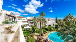 Charming view of Andalusian-style whitewashed buildings and lush gardens surrounding a turquoise swimming pool with palm trees and terracotta paths.